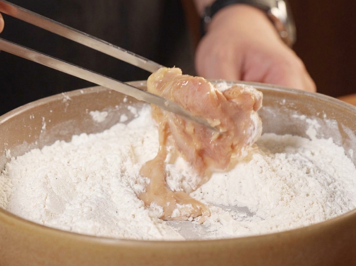 Pinze da cucina che mettono un pezzo di pollo bagnato nel mix di farina secca.