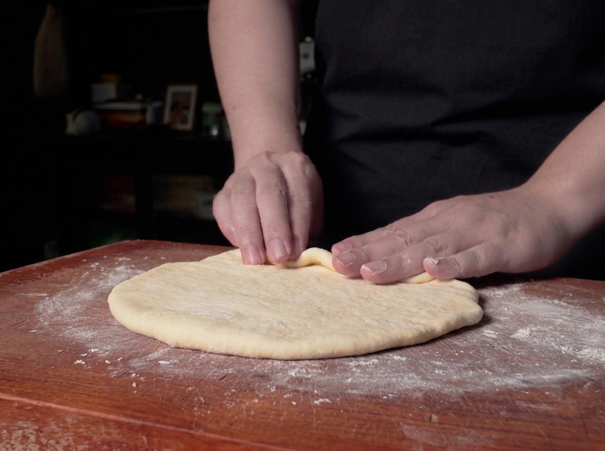 Mani che iniziano ad arrotolare il bordo di un foglio di impasto piatto su un bancone di legno.