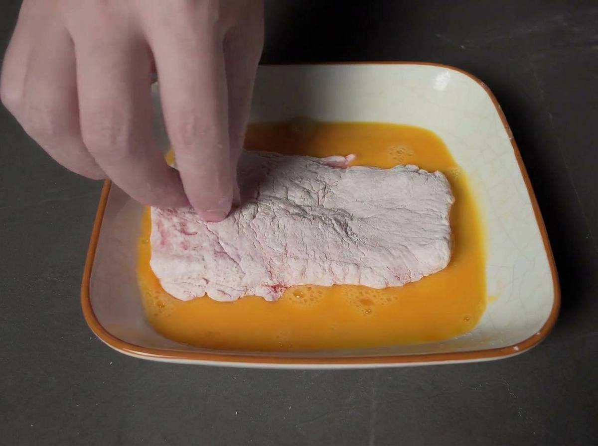 Una mano che preme una cotoletta passata nelluovo in una ciotola piena di pangrattato panko.