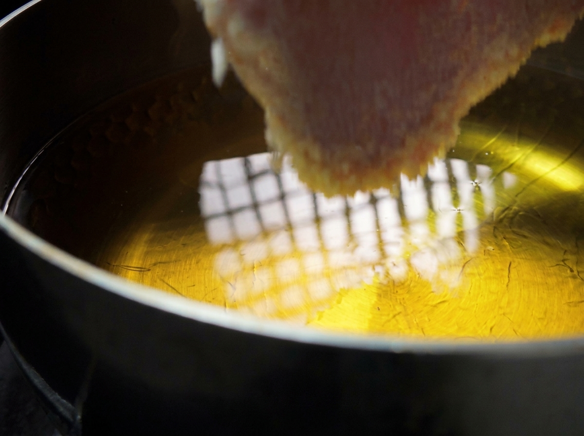 Una spessa cotoletta di maiale impanata che viene immersa con cura in una pentola di olio bollente.