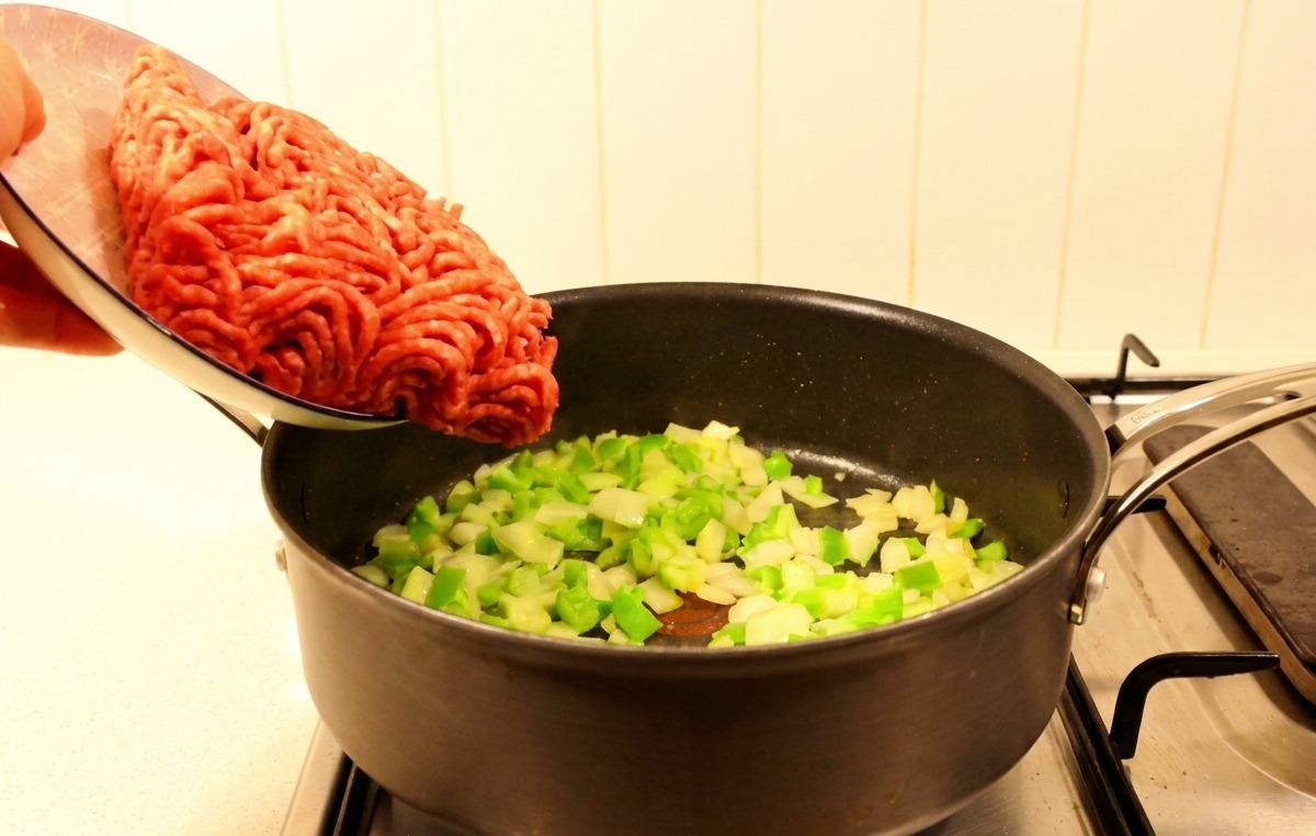 Carne macinata cruda versata da un piatto in una padella con peperoni e cipolle soffritti.