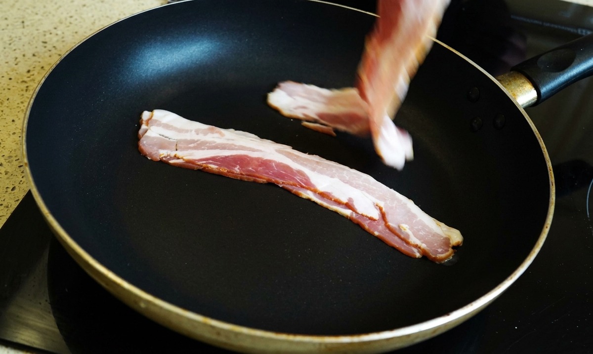 Una mano che adagia fette di pancetta cruda in una padella nera.