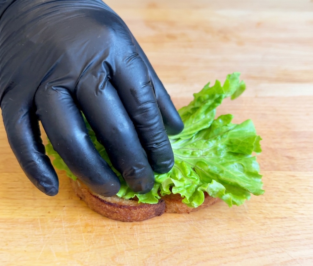 Una mano con un guanto che posiziona un pezzo di lattuga verde brillante ripiegata su una fetta di pane tostato adagiata su un tagliere di legno.