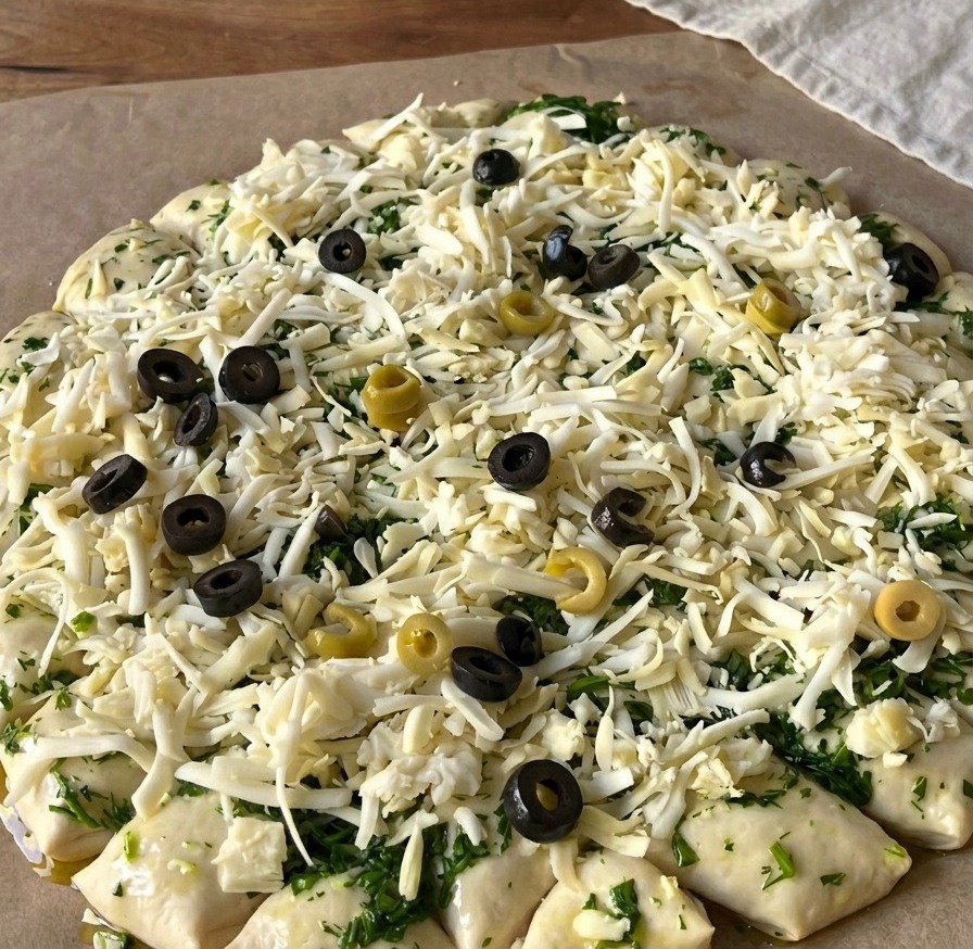 Vista dallalto di un impasto per pizza crudo coperto di formaggio grattugiato e guarnito con numerose rondelle di olive nere e verdi.