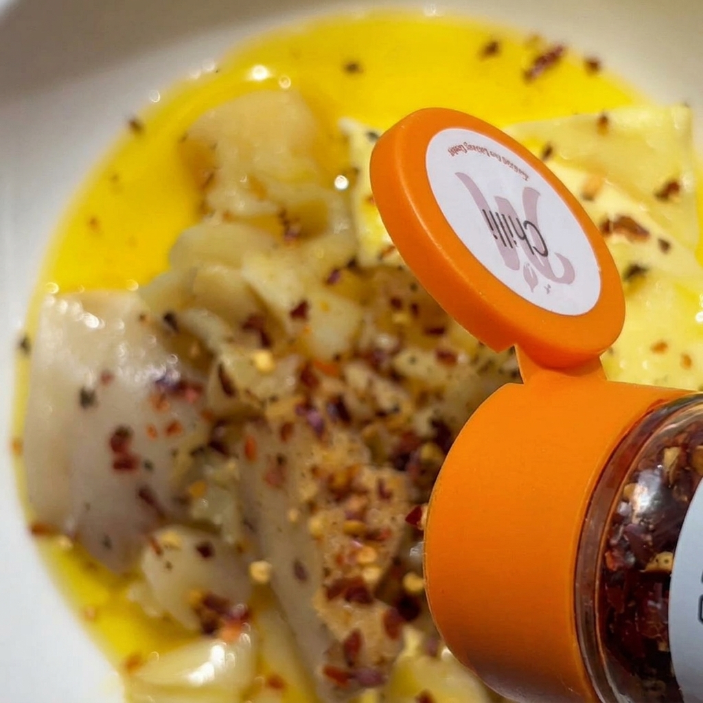 Fiocchi di peperoncino rosso versati da un contenitore per spezie su una ciotola di aglio schiacciato e burro.