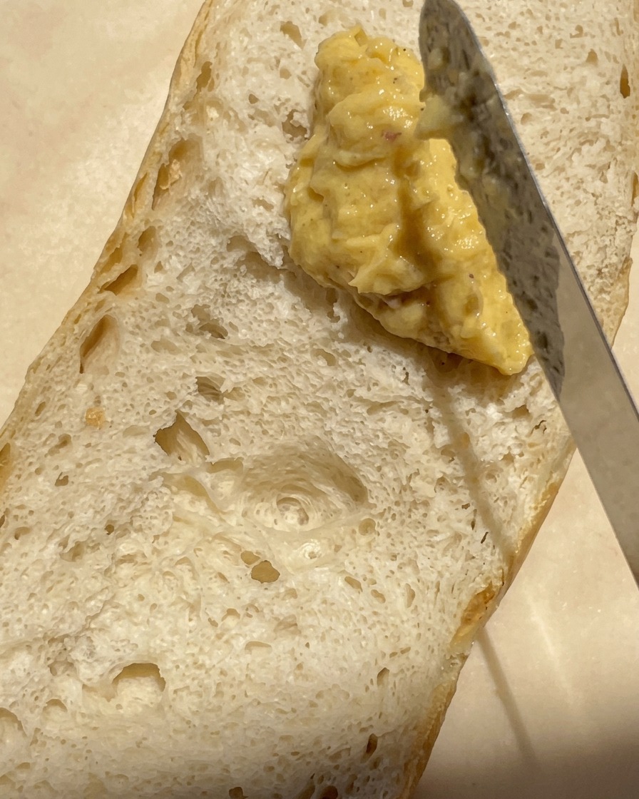Un primo piano di un coltello che spalma una miscela densa e cremosa di aglio arrosto e burro sulla superficie alveolata di mezza baguette.