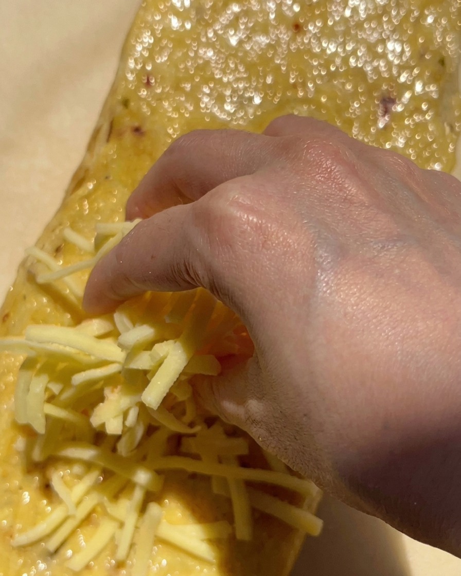 Una mano che distribuisce unabbondante manciata di mozzarella bianca grattugiata su una pagnotta di pane spalmata di burro allaglio.