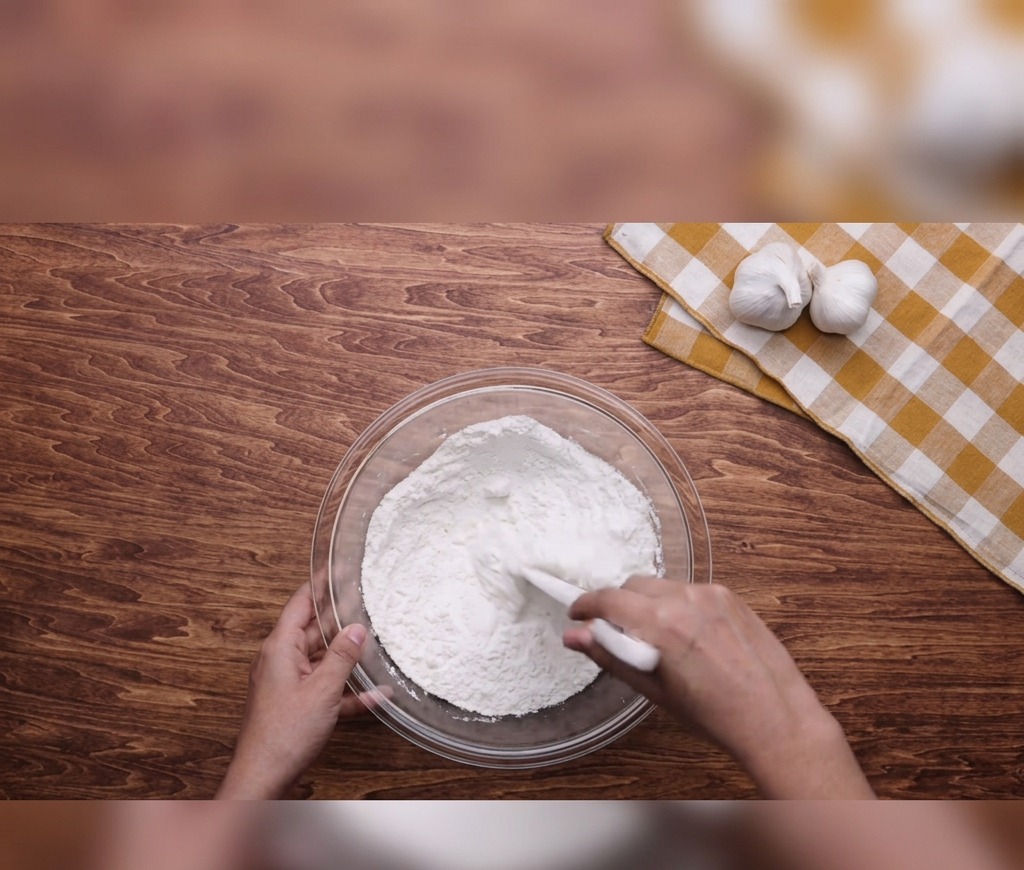 Mani che usano una spatola bianca per mescolare farina, acqua e olio in una ciotola di vetro su un tavolo di legno.