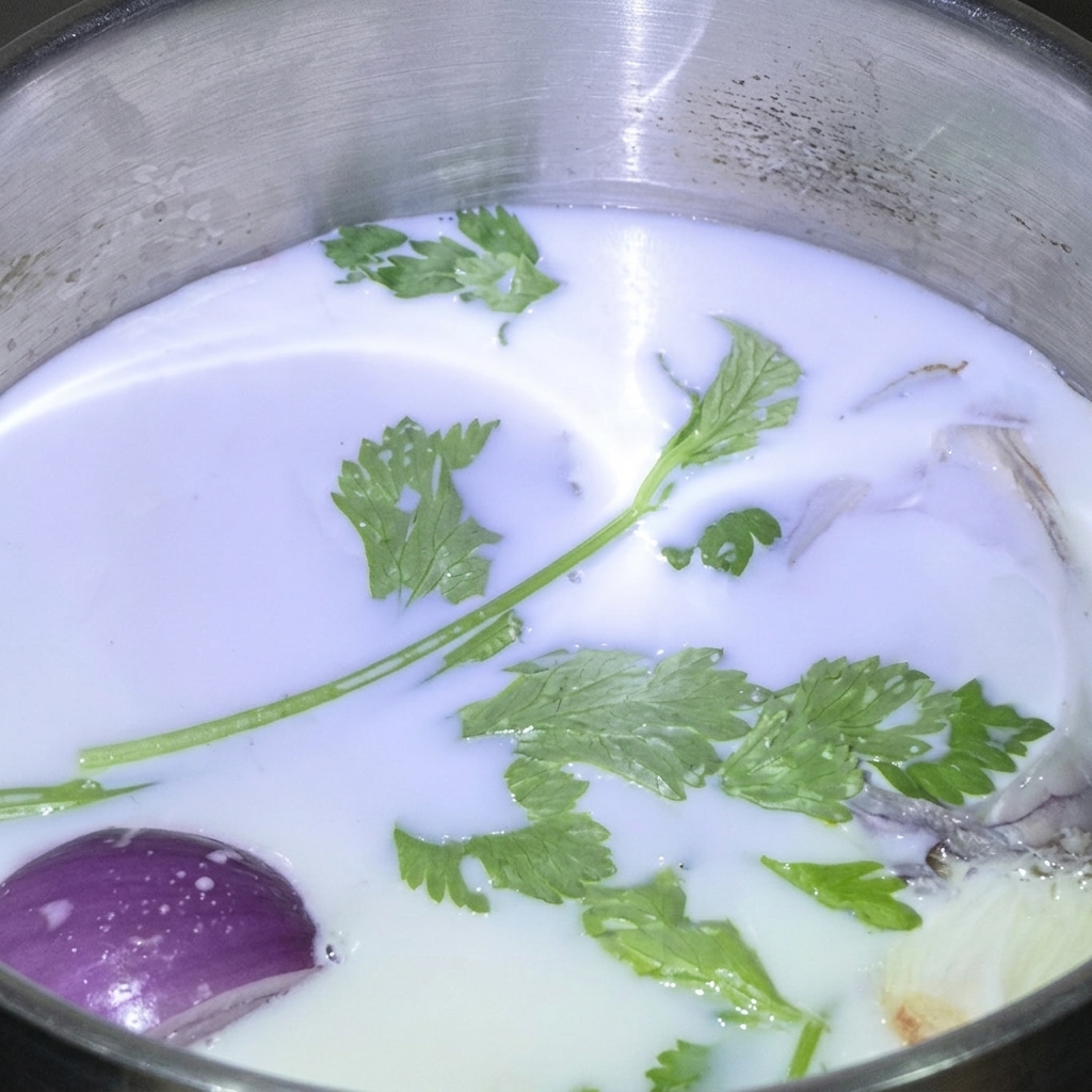 Erbe fresche e uno spicchio di cipolla rossa che galleggiano in una pentola di acciaio inossidabile piena di latte.