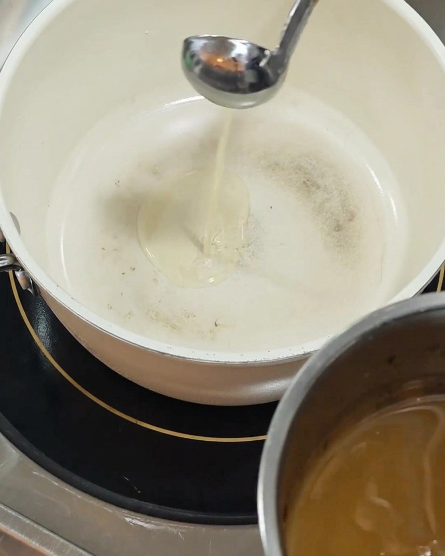 Un mestolo dargento versa dellolio giallo limpido in una pentola di ceramica bianca posizionata su un piano a induzione nero.
