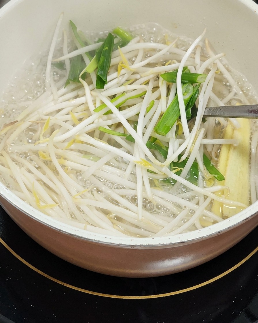 Una pentola con germogli di soia, foglie daglio verde tagliate a pezzetti e pannocchiette di mais che vengono mescolati con un utensile di metallo.