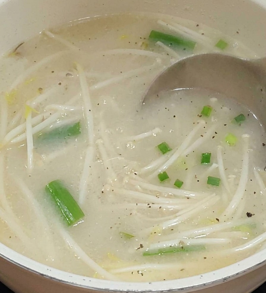 Un mestolo di metallo versa un brodo lattiginoso e chiaro in una pentola contenente germogli di soia e verdure verdi.