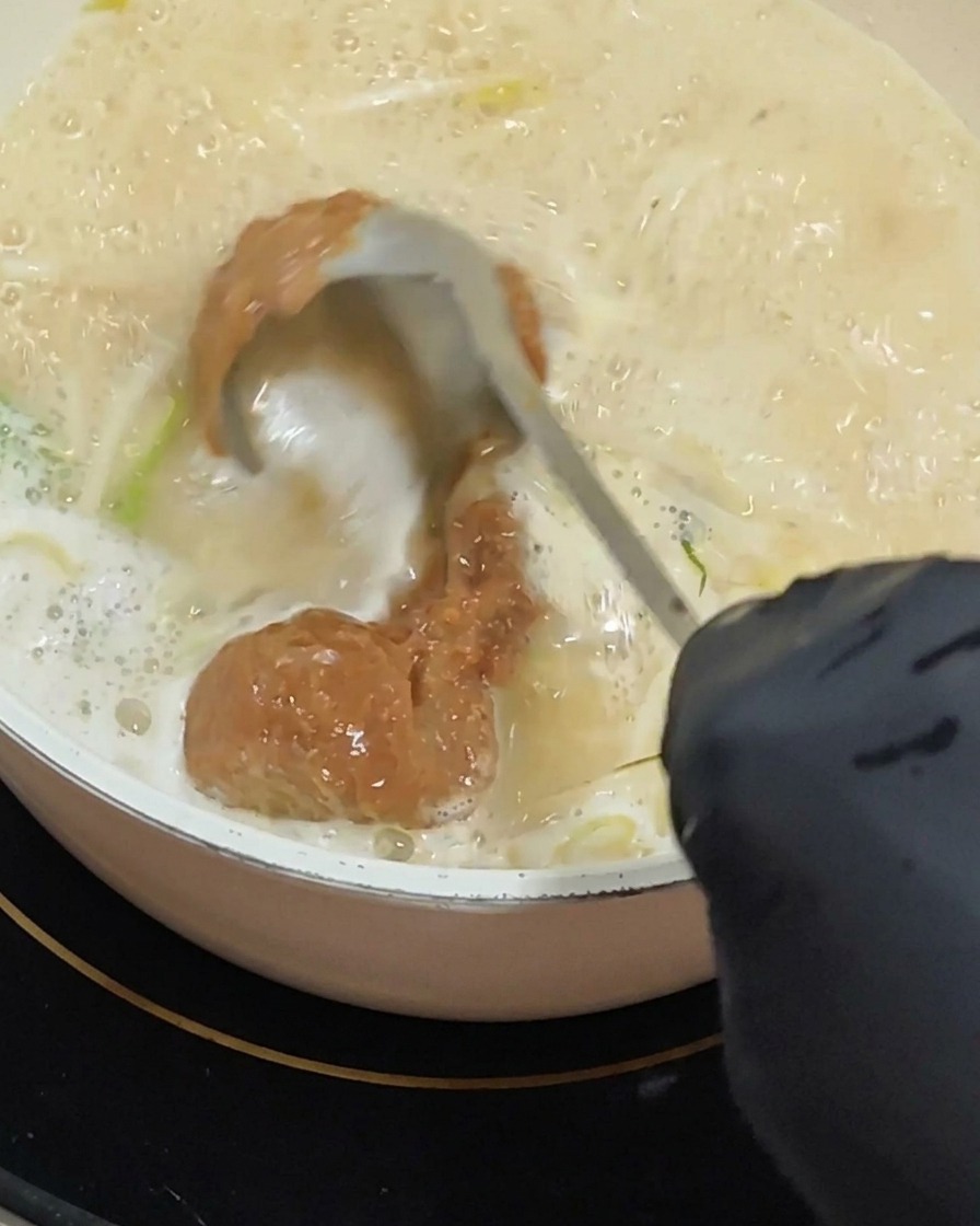 Una mano con un guanto usa un mestolo per stemperare una generosa cucchiaiata di pasta di miso rossastra in una base di zuppa cremosa che sobbolle.