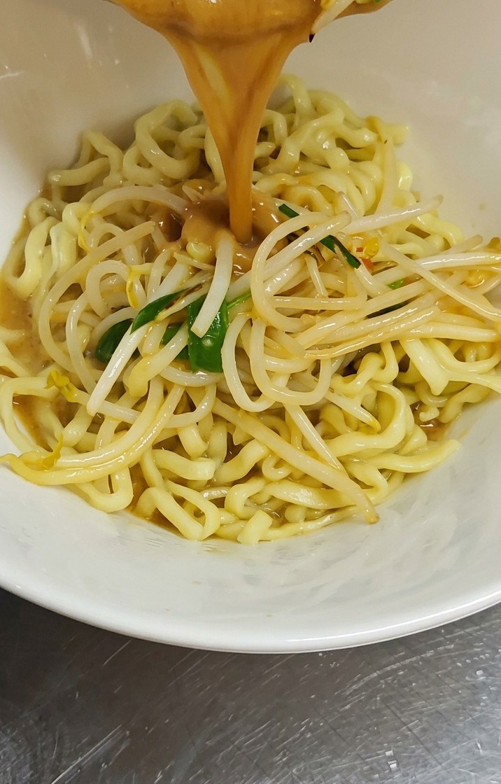 Un flusso di brodo denso al miso marrone e germogli di soia saltati viene versato da una pentola in una ciotola bianca colma di noodle gialli arricciati.