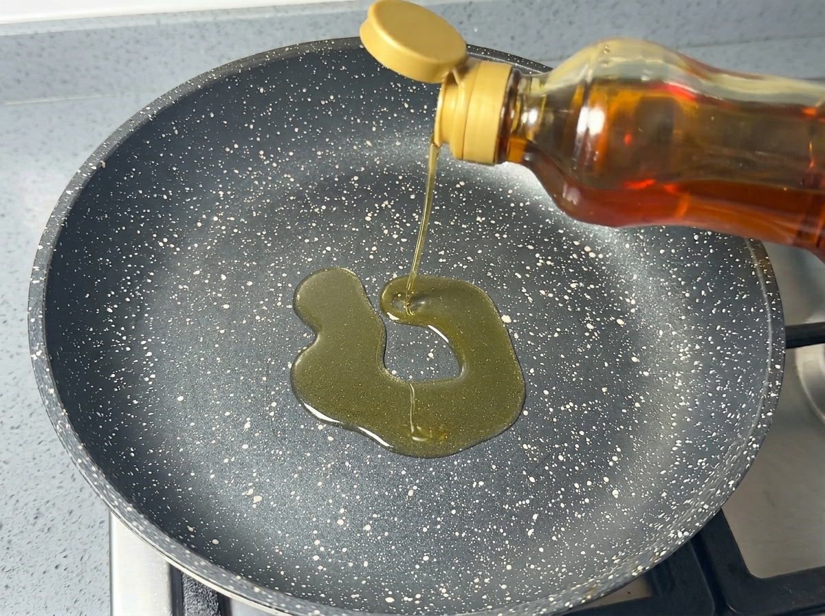 Olio di semi versato da una bottiglia in una padella antiaderente scura e picchiettata.