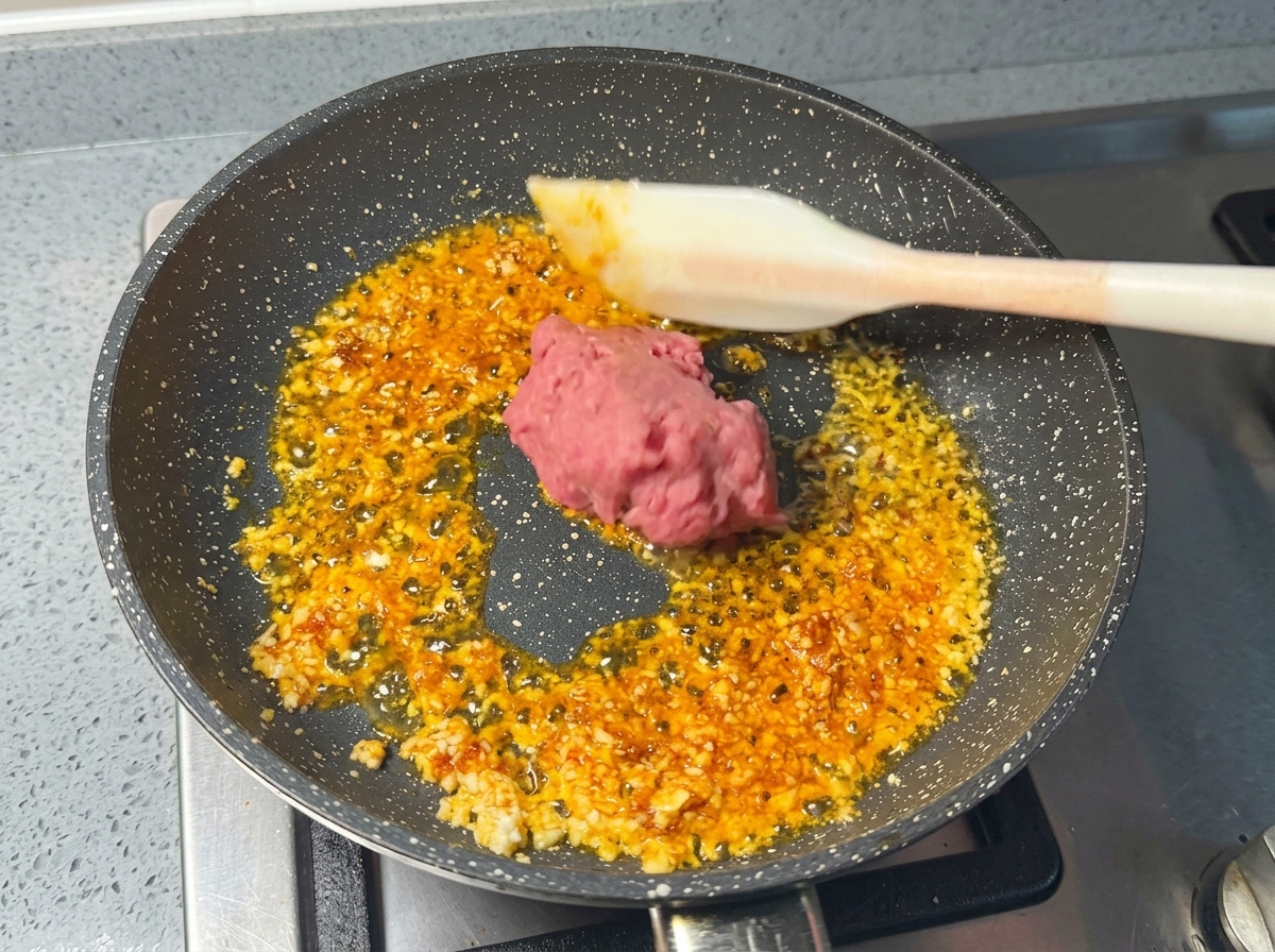 Una porzione di carne di maiale macinata cruda e rosa inserita nella padella con gli aromi usando una spatola di silicone bianca.
