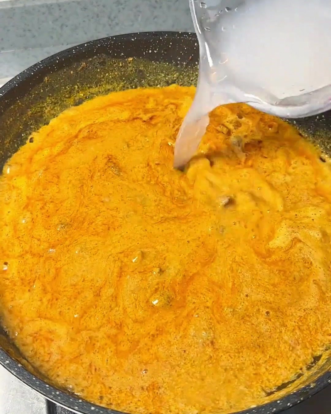 Acqua limpida versata da un contenitore in una miscela di curry arancione e densa che sobbolle in una padella scura.