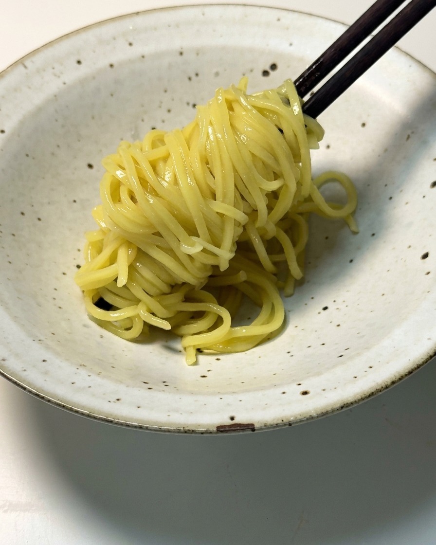 Noodles ramen gialli cotti sollevati con bacchette di legno scuro e adagiati in una ciotola di ceramica chiara.