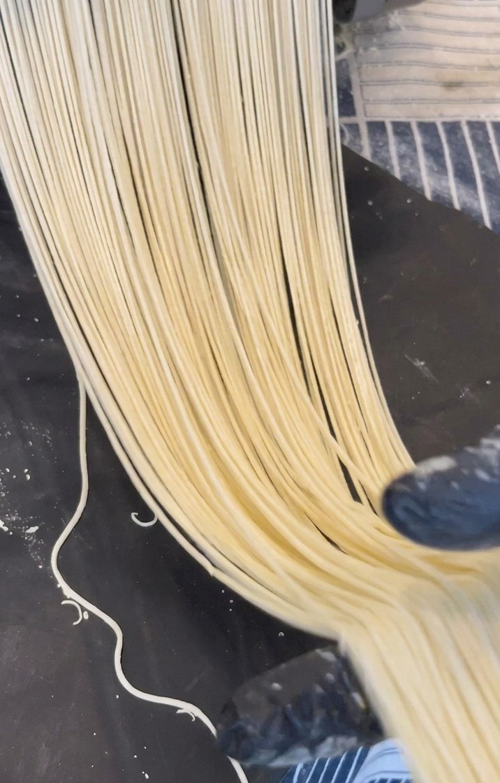 Lunghi fili di noodles per ramen di colore giallo pallido che fuoriescono da una macchina tirapasta in metallo su una superficie scura.