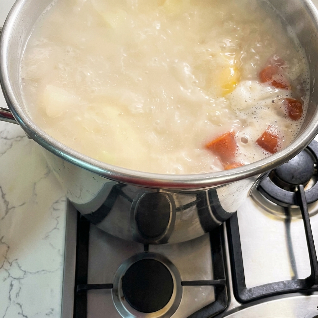 Una grande pentola in acciaio inossidabile su un fornello a gas riempita di brodo bollente, cremoso, di colore bianco e arancione.