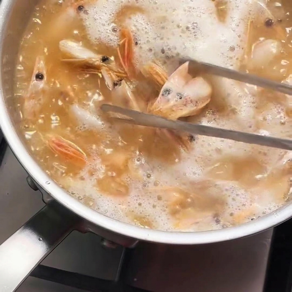 Teste di gambero mescolate con pinze metalliche in una pentola di olio bollente che sta assumendo un colore arancione intenso.