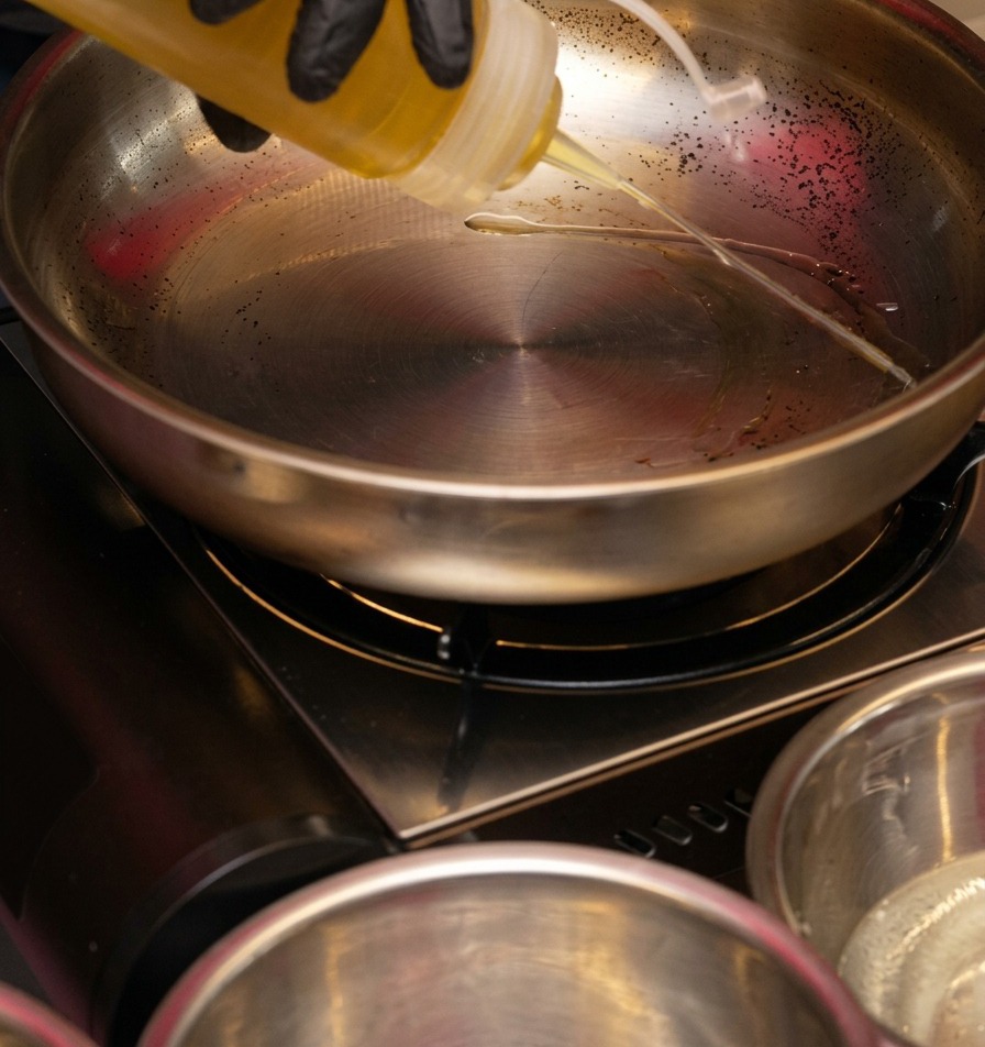 Olio di semi limpido versato da una bottiglia dosatrice in una padella lucida in acciaio inossidabile su un fornello portatile.