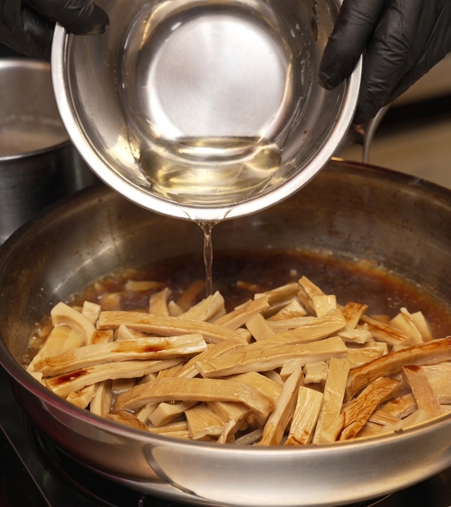 Una persona con guanti neri versa un liquido trasparente da una ciotolina in una padella con germogli di bambù e salsa scura.