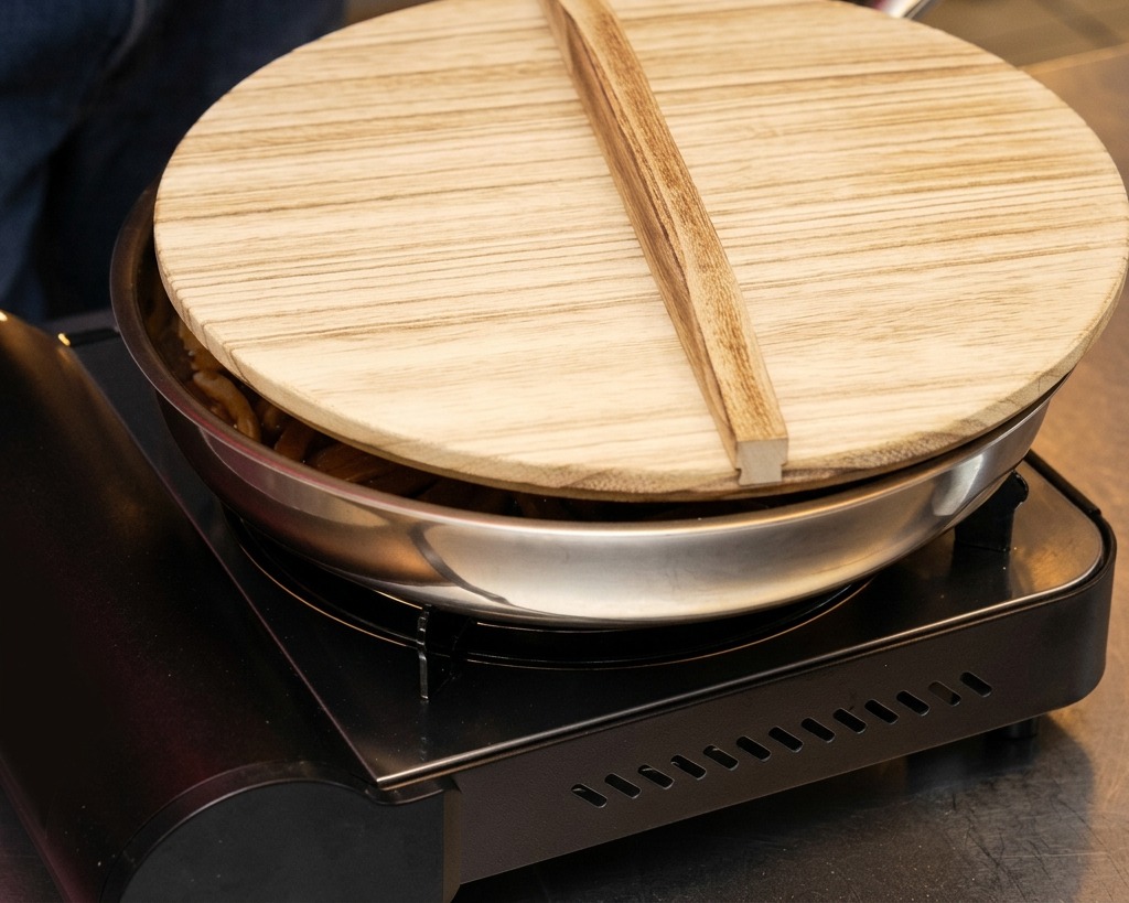 Una padella in acciaio inox con germogli di bambù su un fornello a gas portatile, coperta con un tipico coperchio di legno.