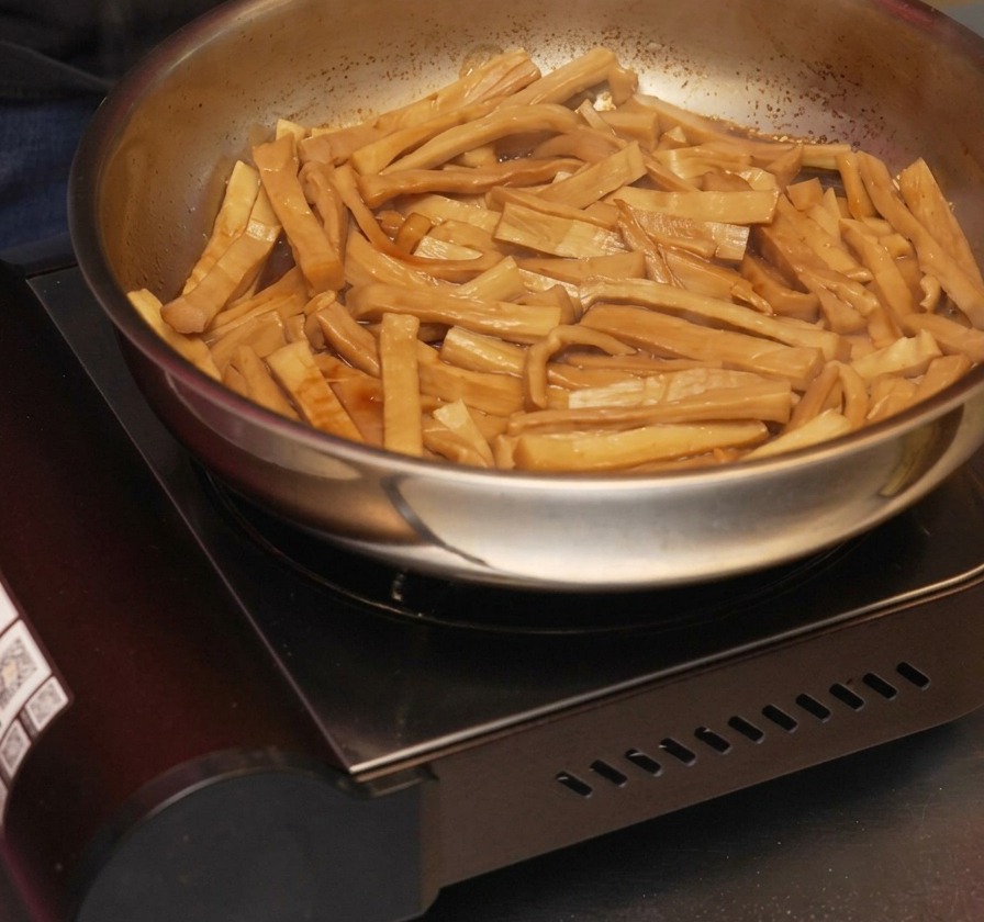 Olio di sesamo versato a filo in una padella di germogli di bambù conditi che saltano in una salsa scura e ristretta.
