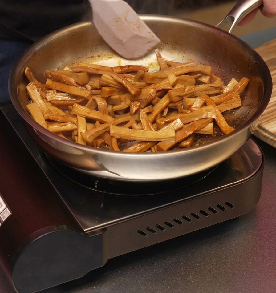 Una spatola bianca muove i germogli di bambù glassati e marroni in padella per distribuire lolio di sesamo aromatico.