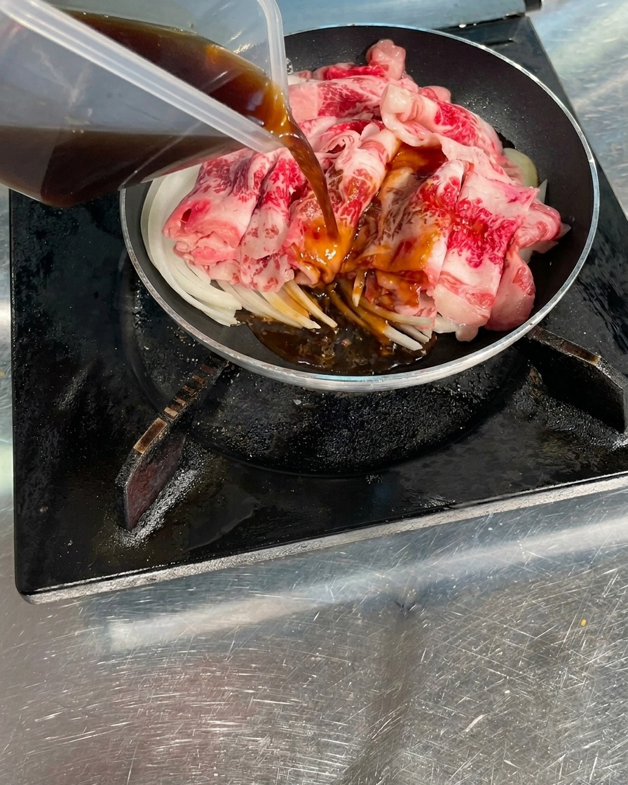 Una salsa saporita di colore marrone scuro che viene versata da un contenitore trasparente sulle fette di manzo crudo e sulle cipolle in padella.