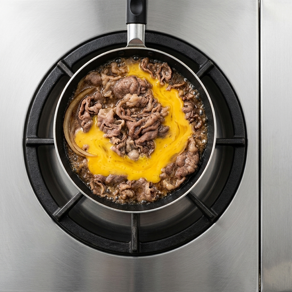 Uovo liquido sbattuto giallo versato sopra le fette di manzo e le cipolle cotte a fuoco lento allinterno di una padella a fondo piatto.