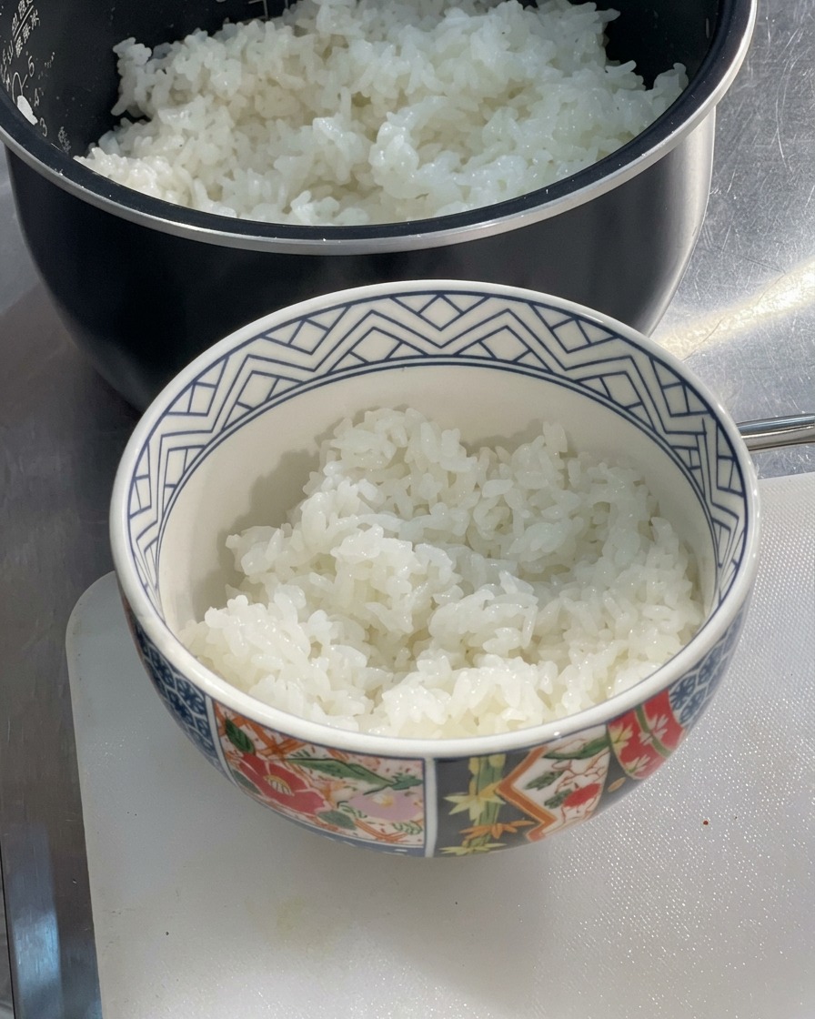 Una ciotola di ceramica decorata riempita a metà con riso bianco al vapore, soffice e appena cotto, appoggiata su un tagliere accanto a un cuociriso.