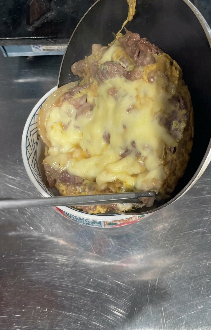 Una padella inclinata sopra una ciotola di riso bianco, con un fitto strato di manzo cotto e formaggio giallo fuso che scivola via per coprire la superficie.
