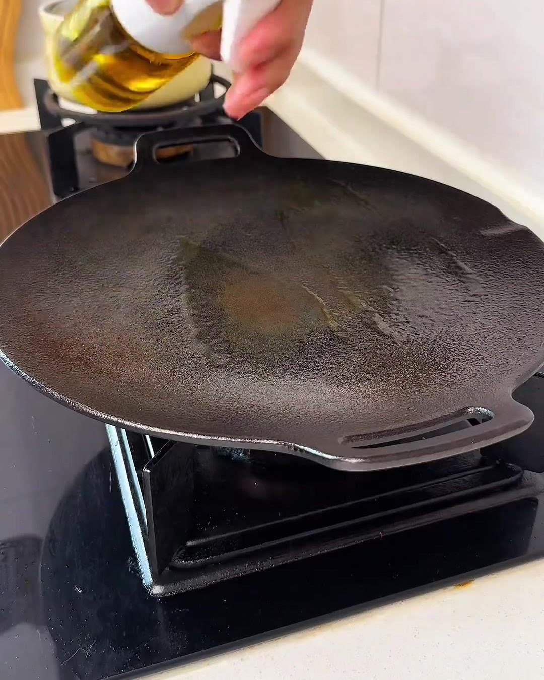 Una mano che spruzza un velo leggero di olio da cucina dorato su una piastra nera circolare pulita sul fornello.