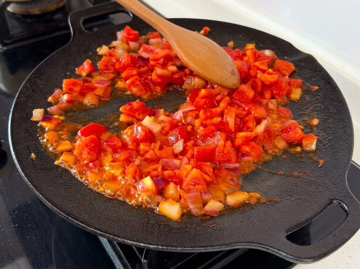Pomodori a cubetti leggermente schiacciati che si mescolano con le cipolle in una padella calda, con i succhi visibili che bollono.