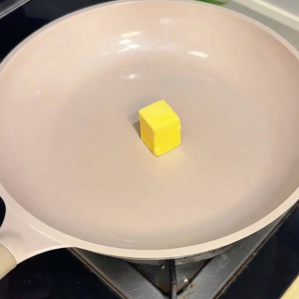 Un cubetto di burro giallo che scivola sulla superficie di una padella antiaderente calda color crema.