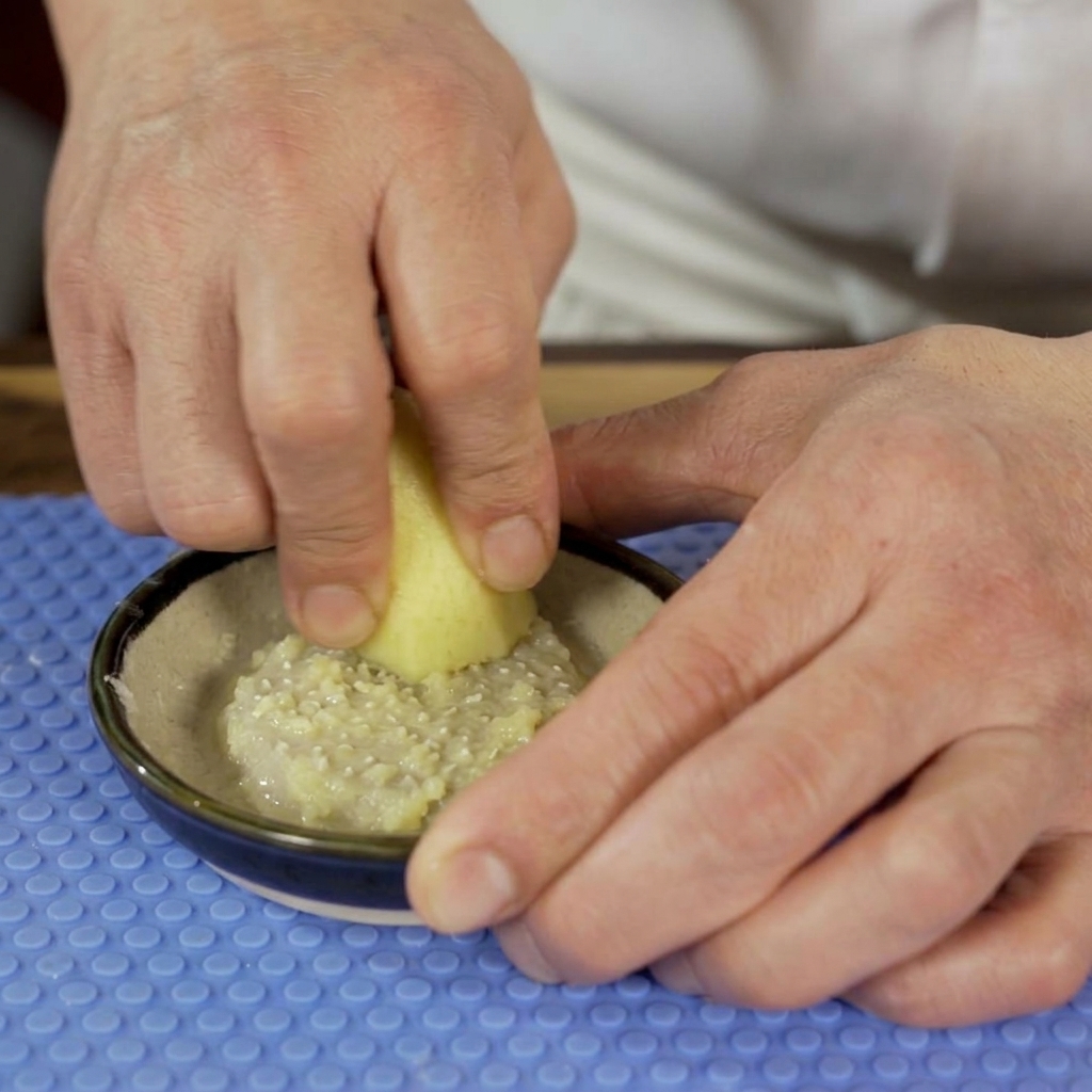 Una persona usa una grattugia in ceramica per ridurre la radice di zenzero fresco in una pasta umida in una ciotolina.