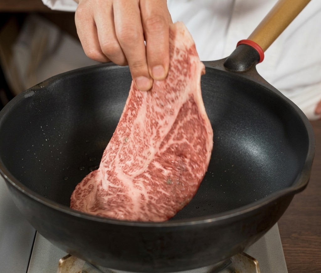 Una mano adagia una grande bistecca cruda ben marezzata e pepata allinterno di una padella in ghisa nera bollente.