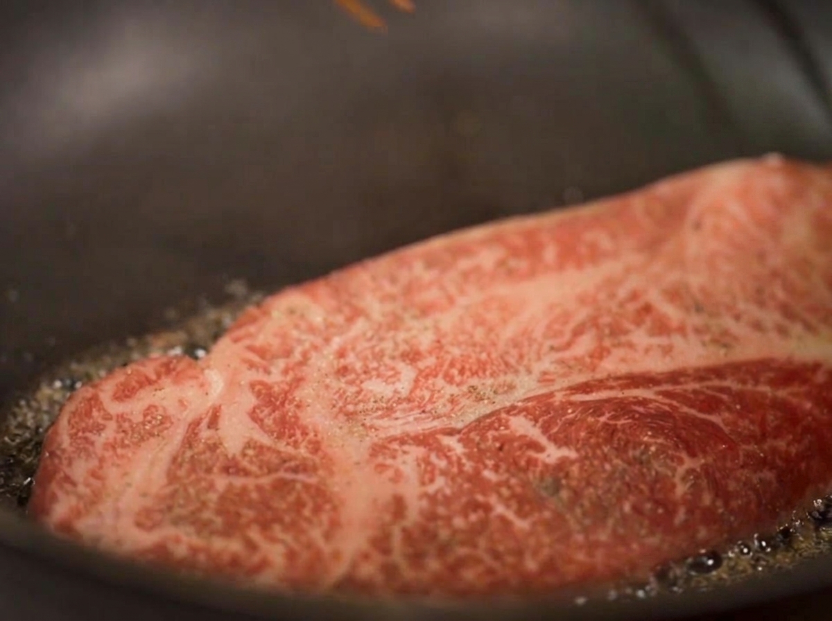 Primo piano di una bistecca spessa che scotta nellolio, con vapore visibile e i bordi inferiori che diventano dorati.