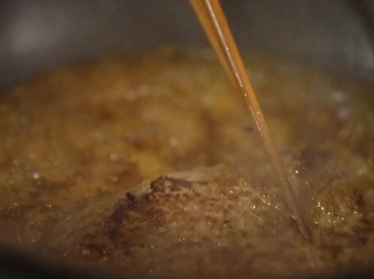 Primo piano di una bistecca che sobbolle in una densa salsa marrone, nappata continuamente con laiuto di bacchette.