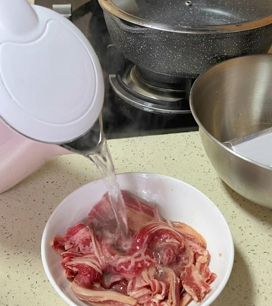 Acqua bollente fumante viene versata da un bollitore elettrico rosa e bianco su straccetti di manzo crudo in una ciotola.