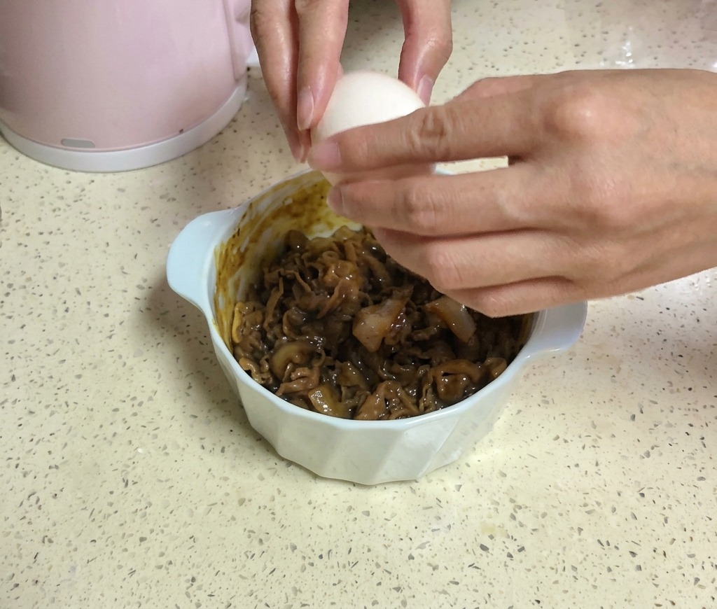 Due mani rompono un uovo bianco sopra una ciotola di manzo guarnita con due fette di formaggio giallo.