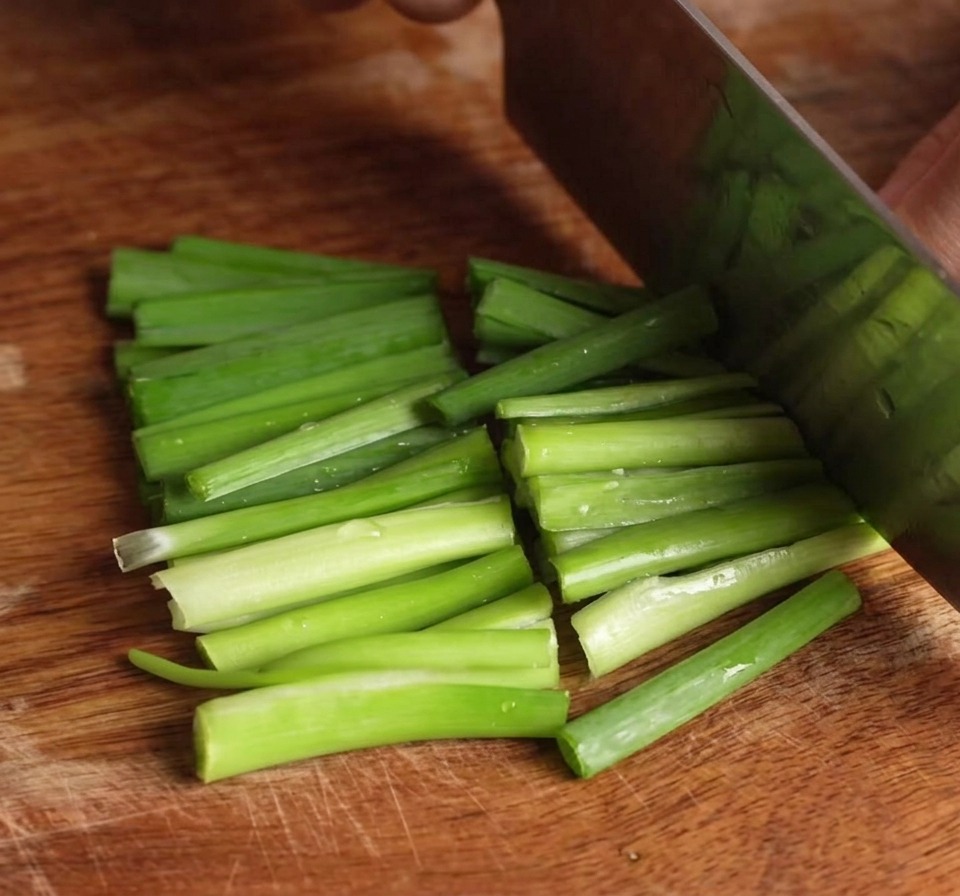 Un coltello che taglia un mazzetto di erba cipollina verde brillante su un tagliere di legno creando piccoli segmenti.
