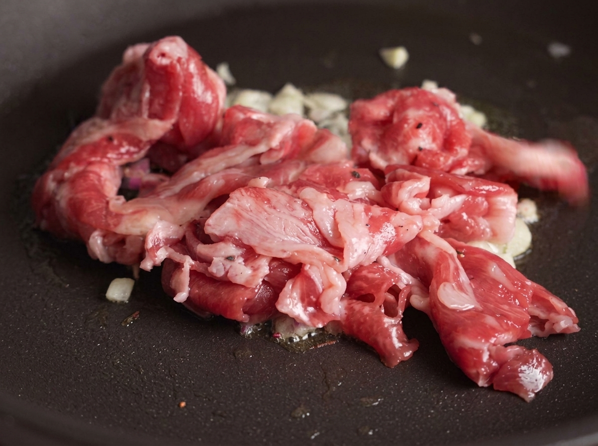 Fette di manzo crudo aggiunte a una padella con aglio soffritto con alcune parti della carne che iniziano a dorarsi per il calore.