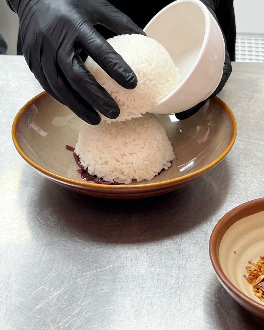 Mani con guanti che sollevano una piccola ciotola bianca per mostrare una cupola di riso cotto a vapore appoggiata sulle cipolle rosse.