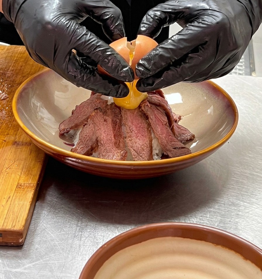 Due mani protette da guanti che rompono delicatamente un uovo onsen sulla cima di una montagna di carne a fette.