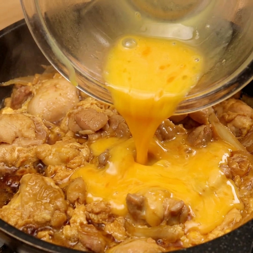 Uova sbattute dorate versate da una ciotola di vetro in una padella con pollo e cipolle che sobbollono in una salsa scura.