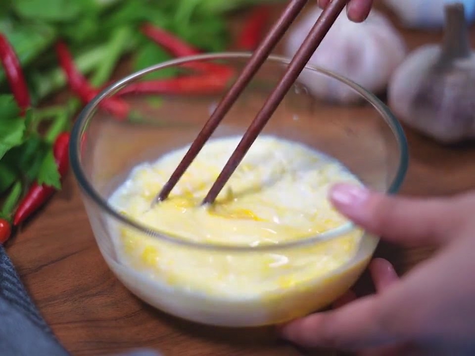 Un paio di bacchette di legno scuro che mescolano un composto giallo chiaro di uova, latte e formaggio in una ciotola di vetro.