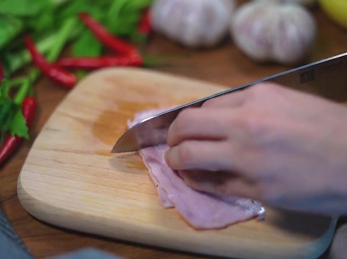 Primo piano di mani che usano un coltello in acciaio inossidabile per tagliare a cubetti strisce di bacon crudo su un tagliere di legno chiaro.